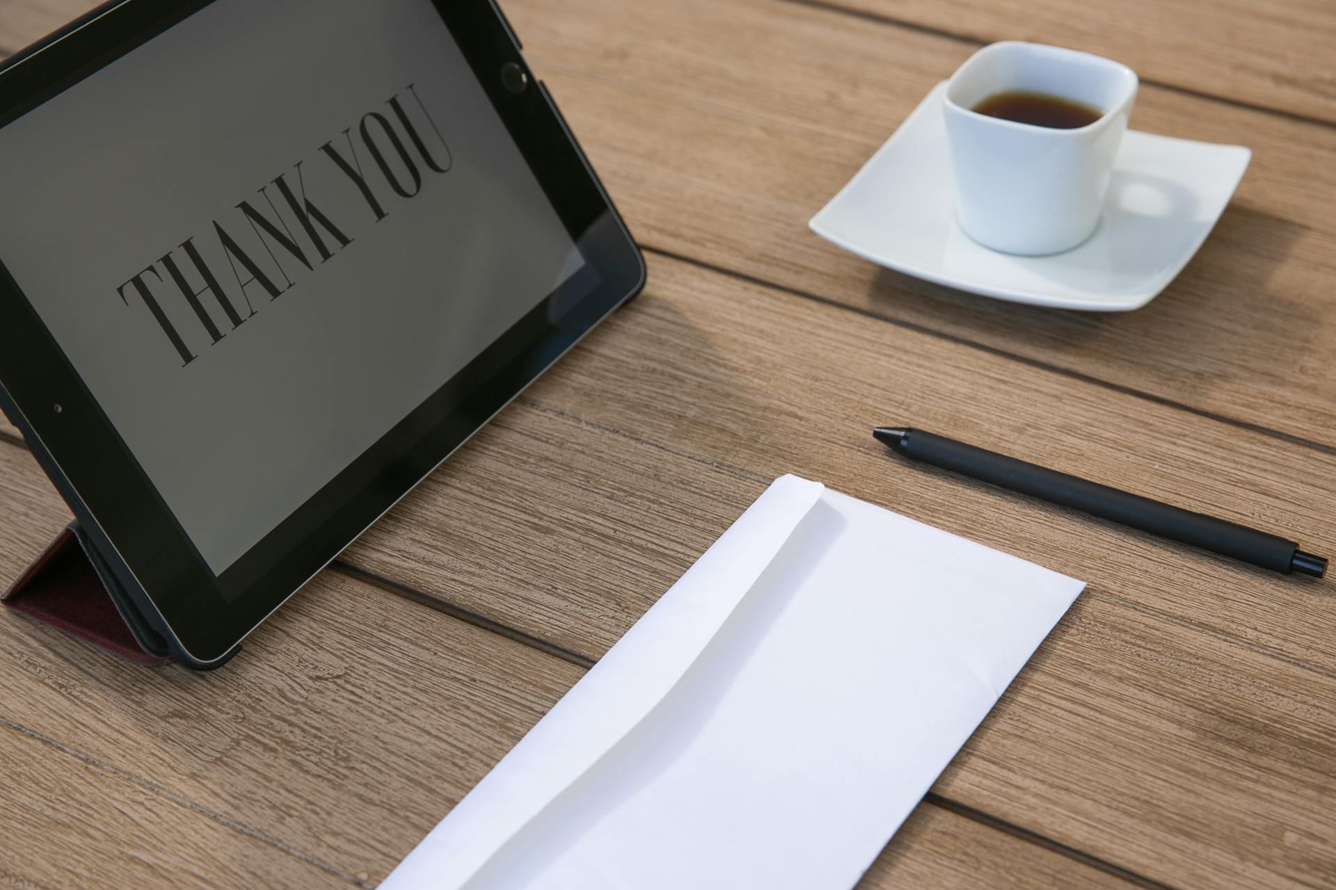 Sophisticated workspace setup with tablet displaying 'Thank You', cup of tea, pen, and envelope on wooden table.