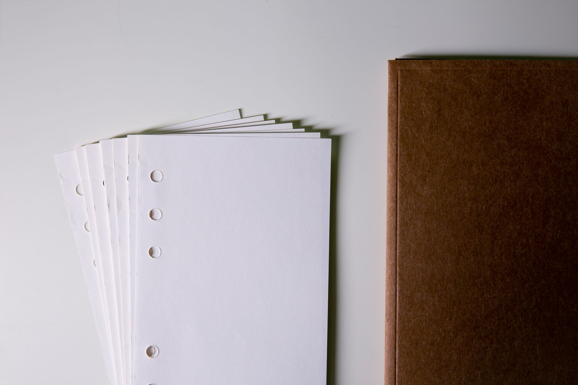 Stack of blank, hole-punched paper next to a closed brown notebook on a white surface.