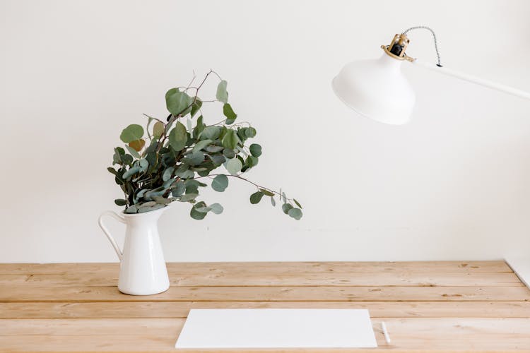 Blank Paper Sheet On A Wooden Desk Under A White Lamp And A Vase With Branches 