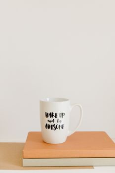 White coffee mug on books with motivational quote 'Wake Up and Be Awesome' on a beige background.
