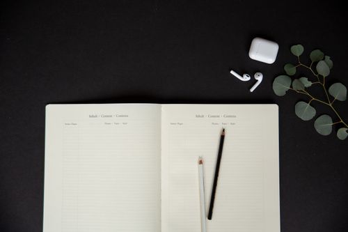 Free A flat lay of an open notebook with pencils, earbuds, and leaves on a dark background. Stock Photo