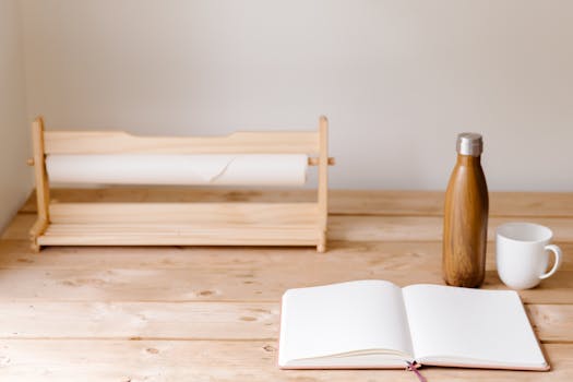 A clean wooden desk featuring an open notebook, tumbler, mug, and paper roll holder. Perfect minimal setup.