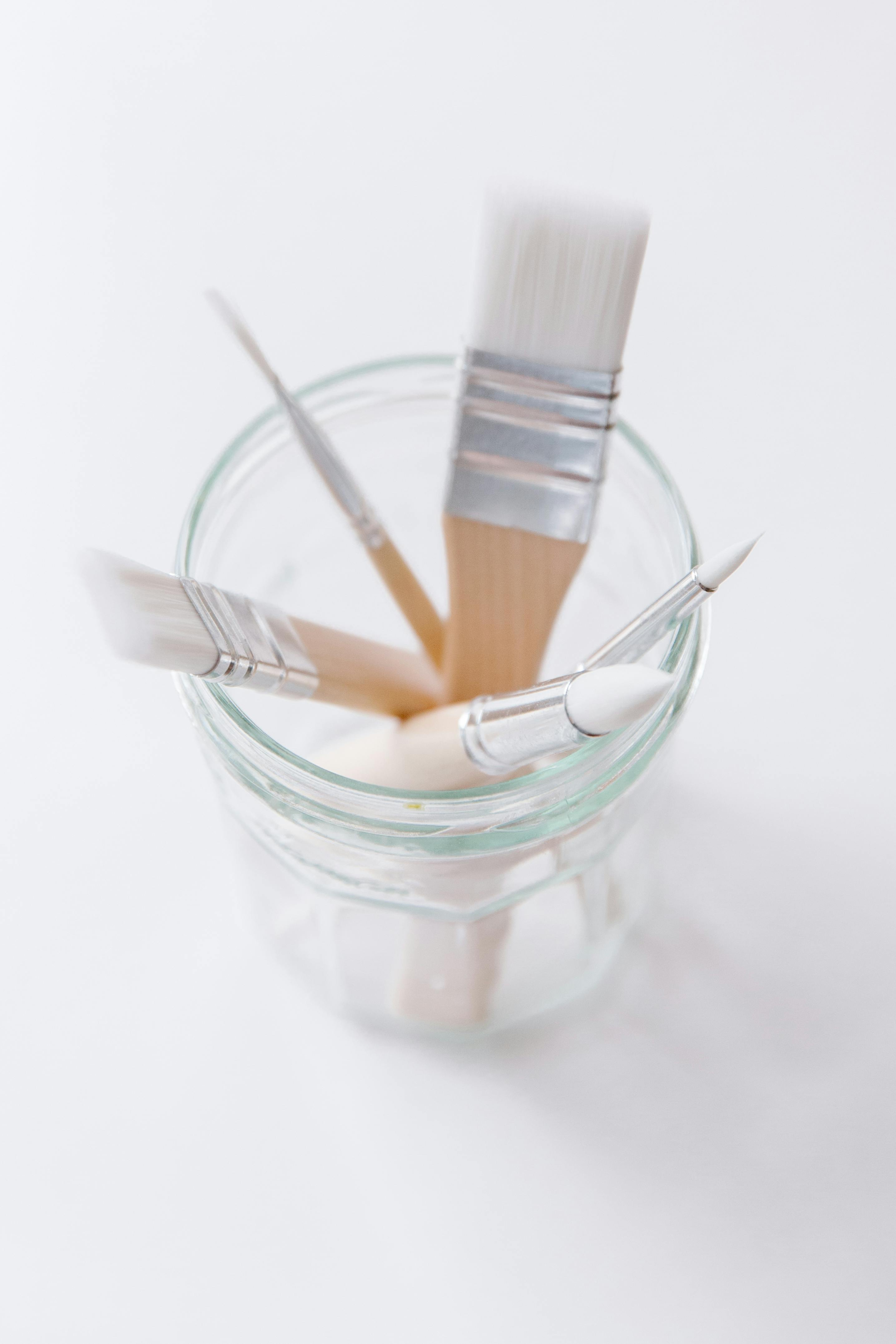 A clean, minimalist close-up of paintbrushes in a glass jar on a white background.