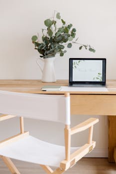 Stylish home office desk scene featuring a laptop, vase with leaves, and a modern chair.