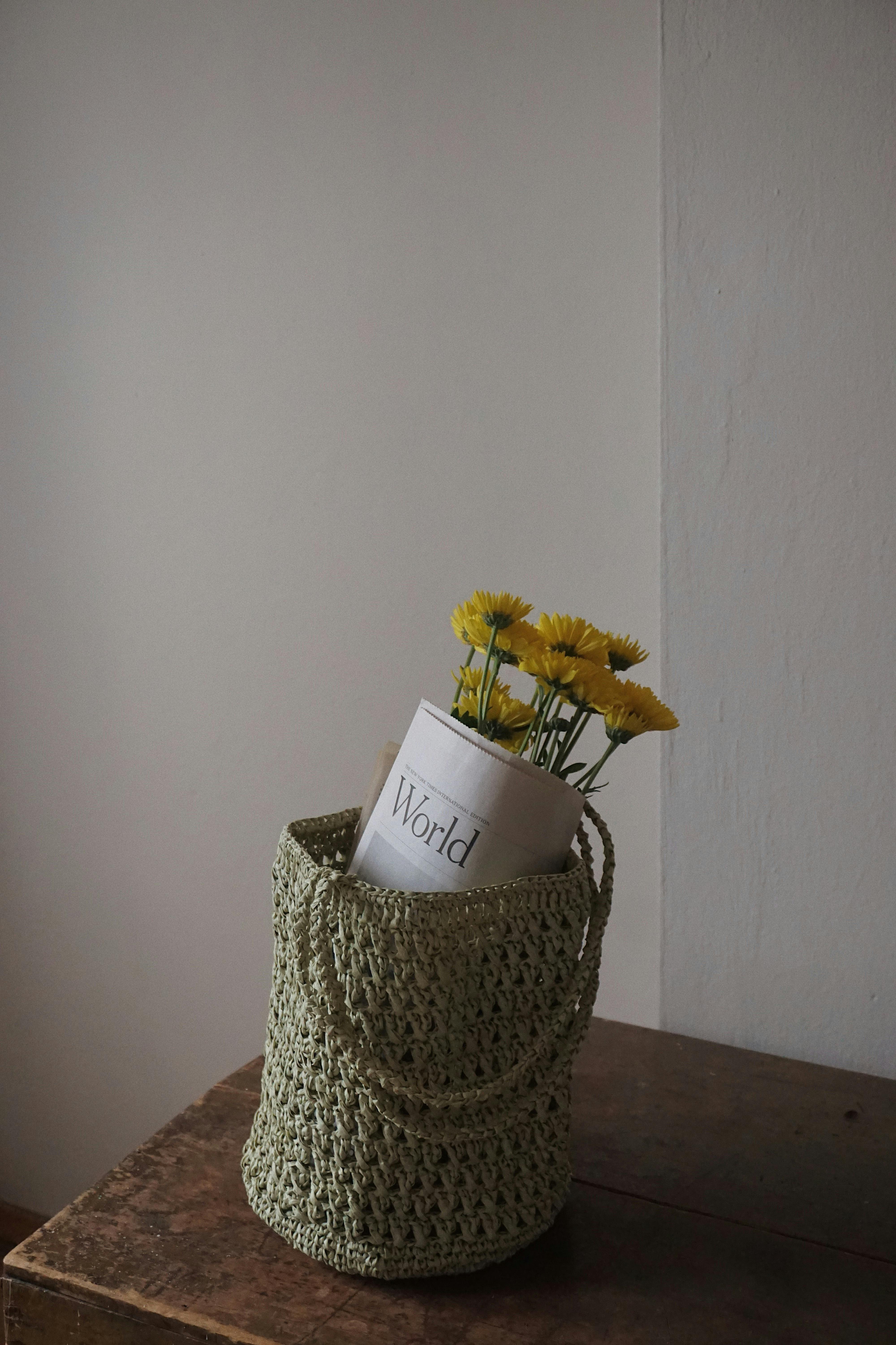 Photograph of a woven green basket containing yellow daisies and a newspaper, set indoors.