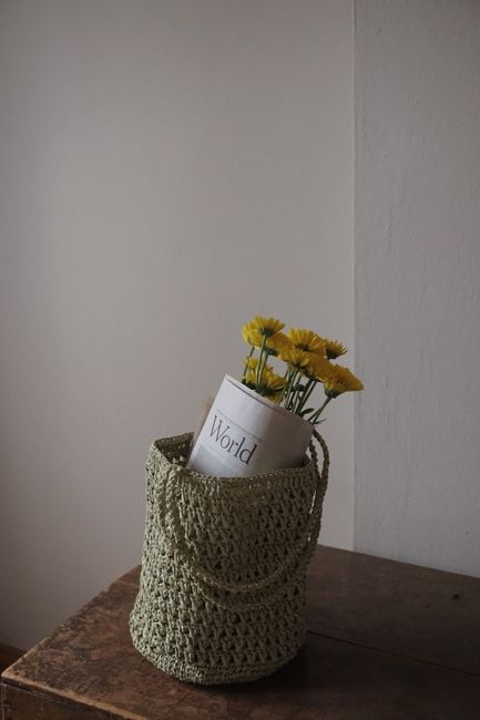 Photograph of a woven green basket containing yellow daisies and a newspaper, set indoors.