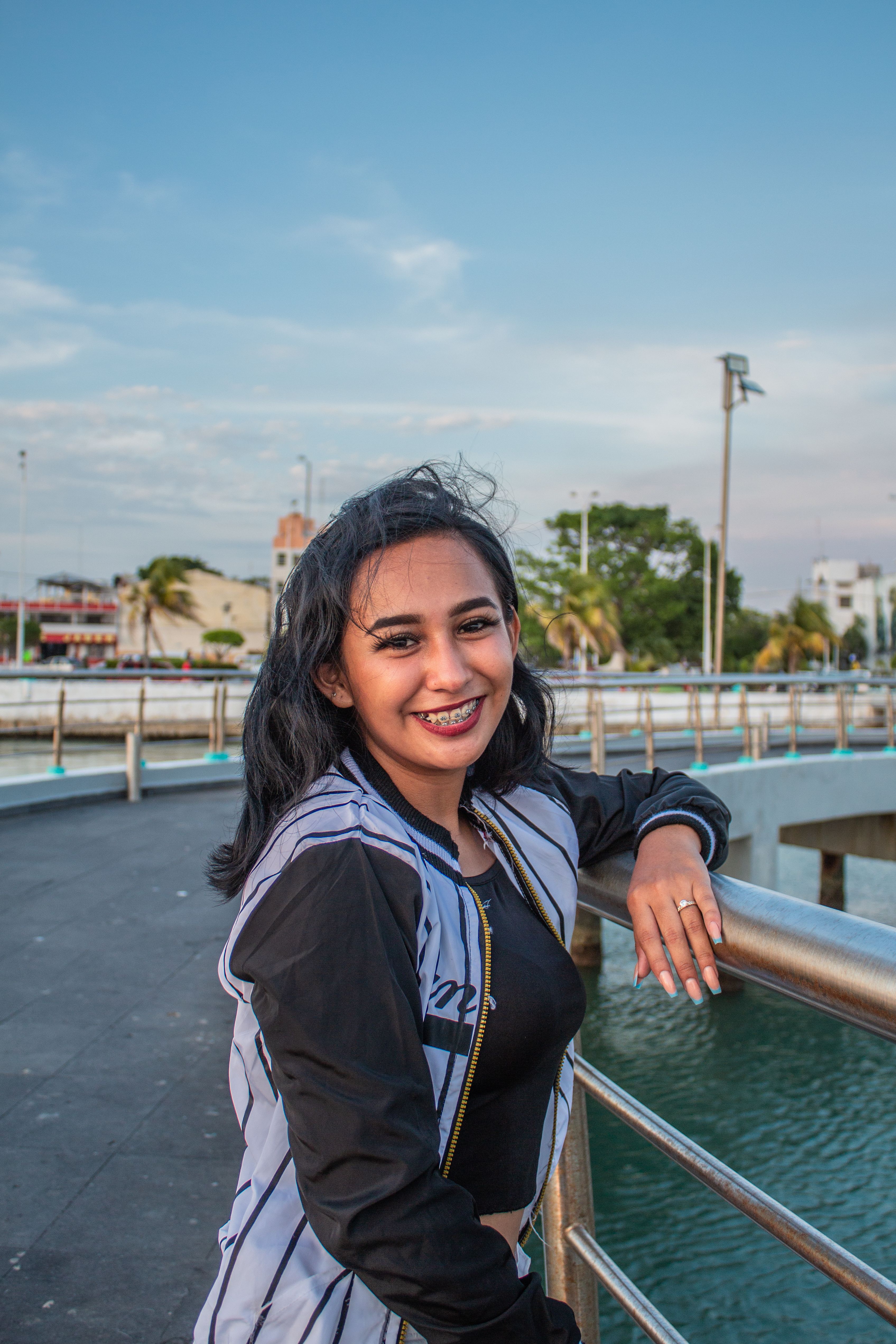 A young woman smiles confidently while leaning on railings at a scenic waterfront under a clear blue sky.