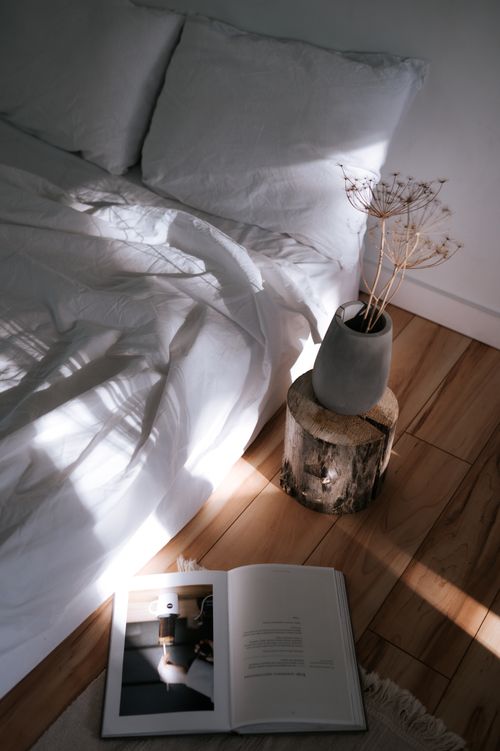 Free A calming minimalist bedroom with sunlight, vase, and open book on a wooden floor. Stock Photo