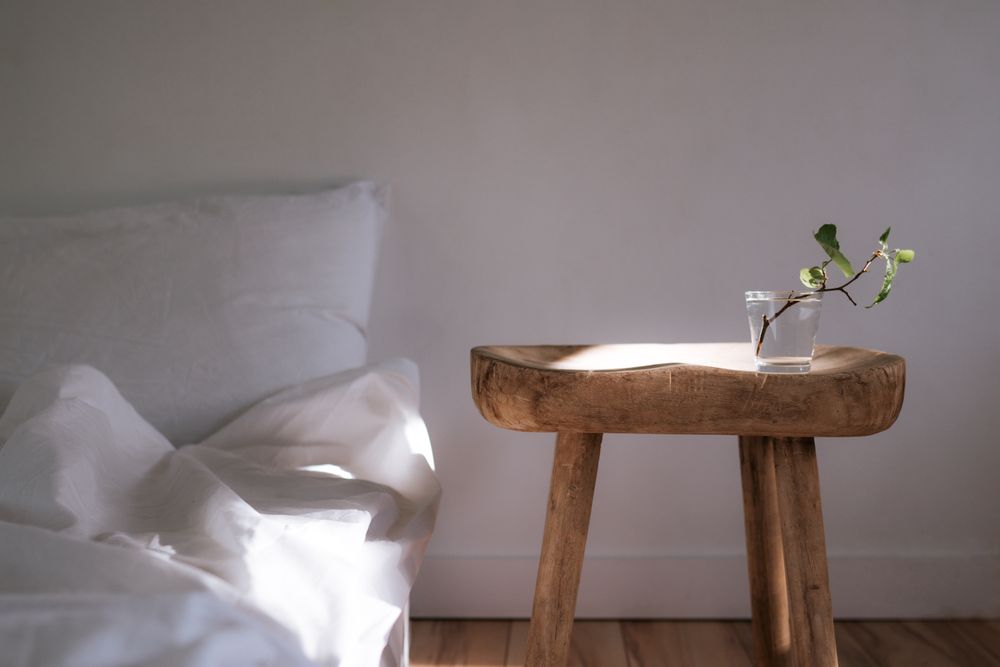 A wooden stool with a single stem in a vase beside an unmade linen bed