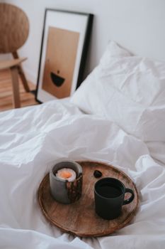 Warm bedroom scene with candle, ceramic mug, and wooden tray on a white blanket, creating a cozy ambiance.