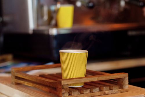 Free Close-up of a steaming yellow paper cup on a wooden tray in a cozy cafe setting. Stock Photo