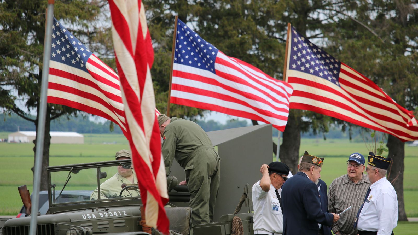 Group of veterans and soldiers by military vehicle with American flags in the background, symbolizing patriotism.