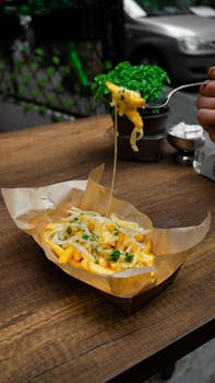Close-up of cheesy french fries being lifted with a fork, served on a wooden table outdoors.
