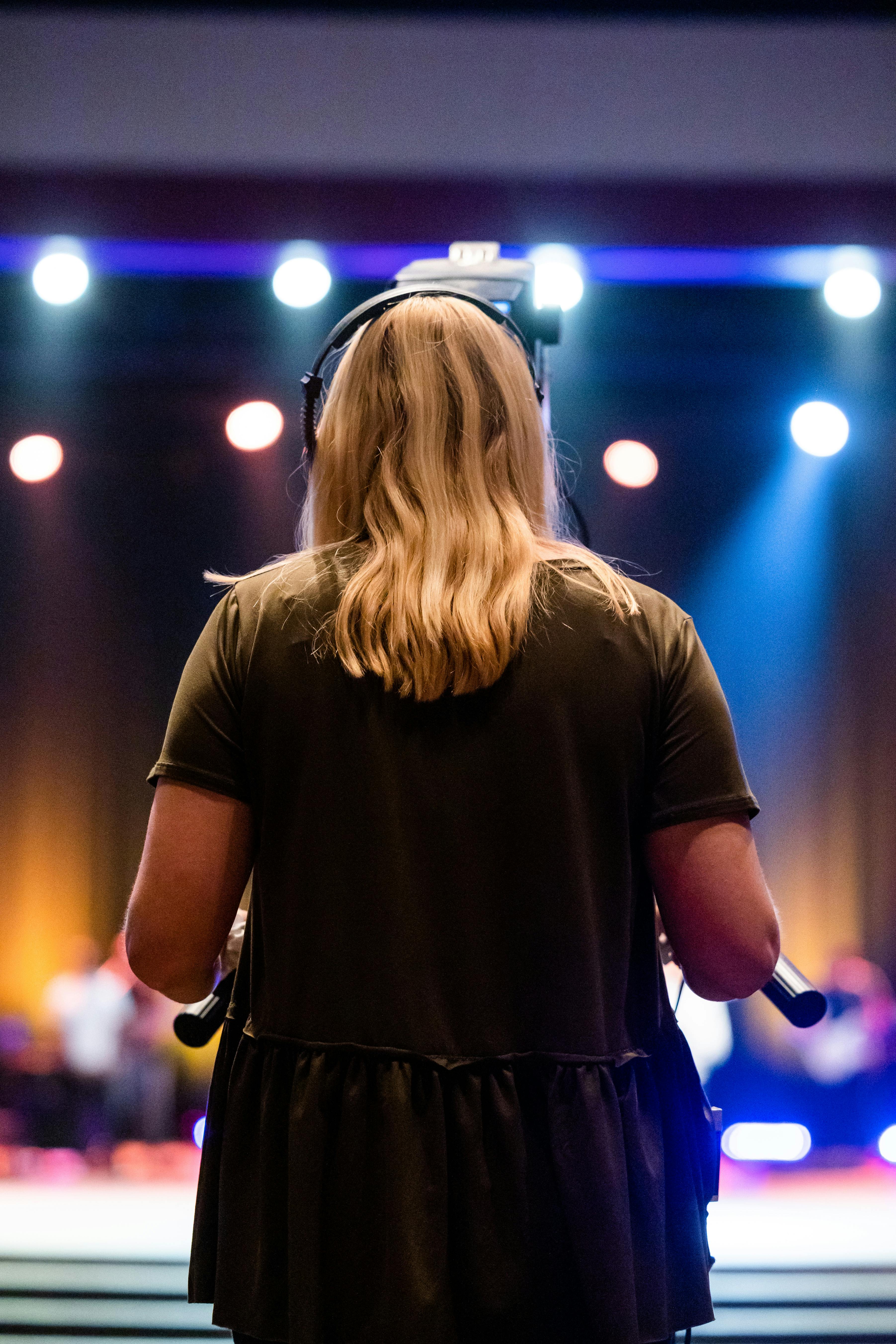 Free A female videographer with headphones captures an event indoors, focused on a stage. Stock Photo