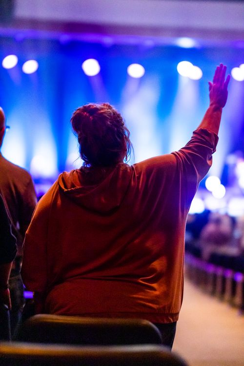 Free A woman in a hoodie raises her hand in a vibrant concert setting with colorful stage lights. Stock Photo