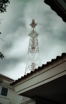 A tall communication tower rises against a cloudy sky with building rooftops in the foreground.