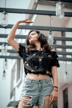 Stylish young woman in ripped shorts and floral top posing outdoors under cafe string lights.