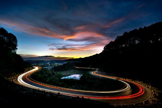 Capture of a winding road at twilight with vibrant light trails under a colorful sky.