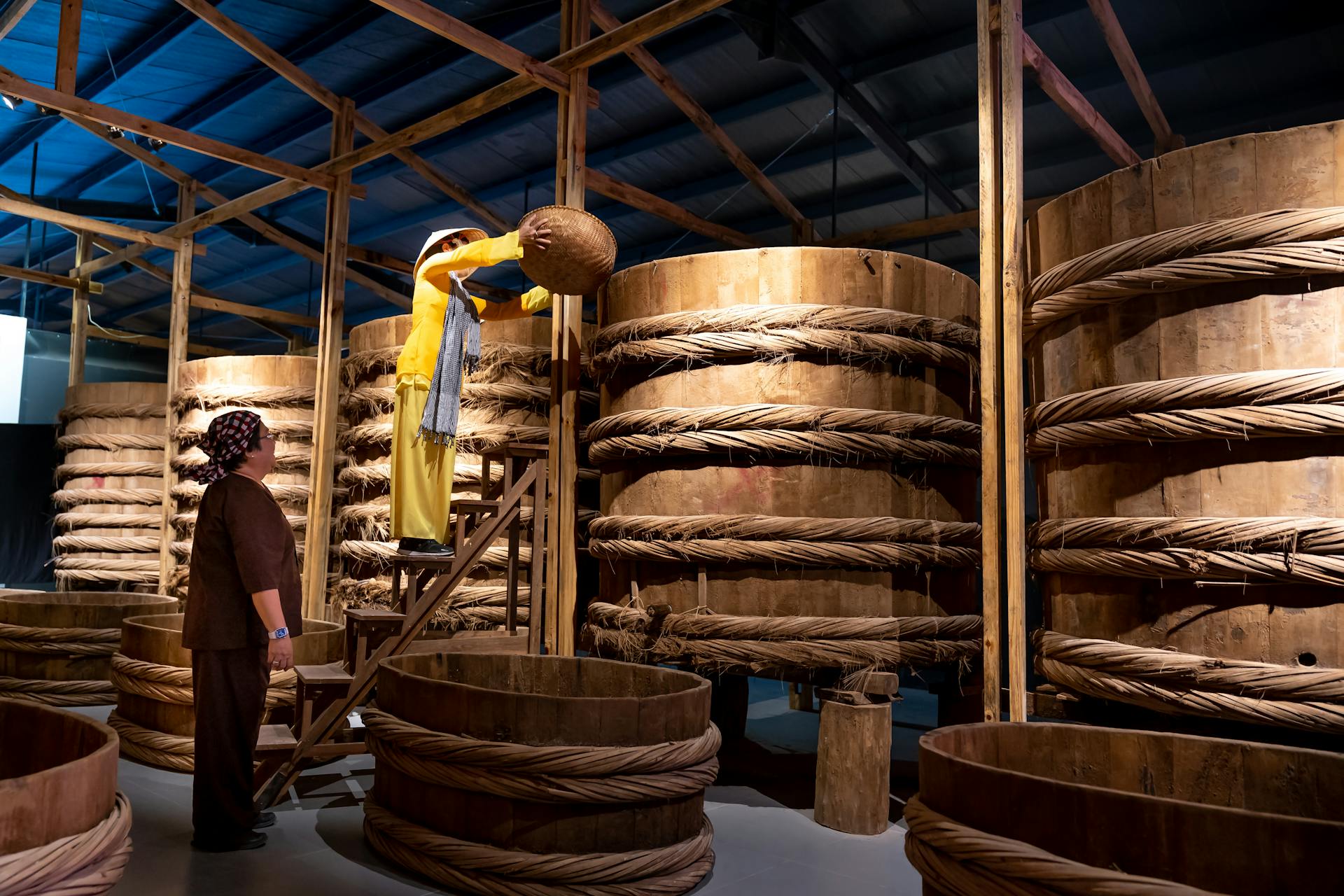 Workers producing traditional fish sauce by pouring into large wooden barrels in an indoor facility.