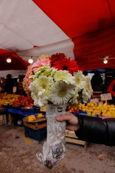 A colorful bouquet of flowers being held at an outdoor market with fresh produce.