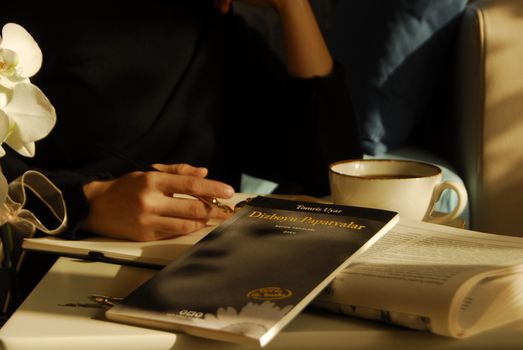 A person enjoys reading with coffee, holding a pen, surrounded by books in warm indoor lighting.