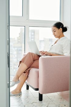 Young woman working from home on a laptop, sitting in an armchair near a large window.