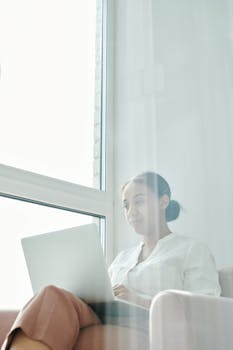 A focused young woman working on her laptop by the window in a bright indoor setting.