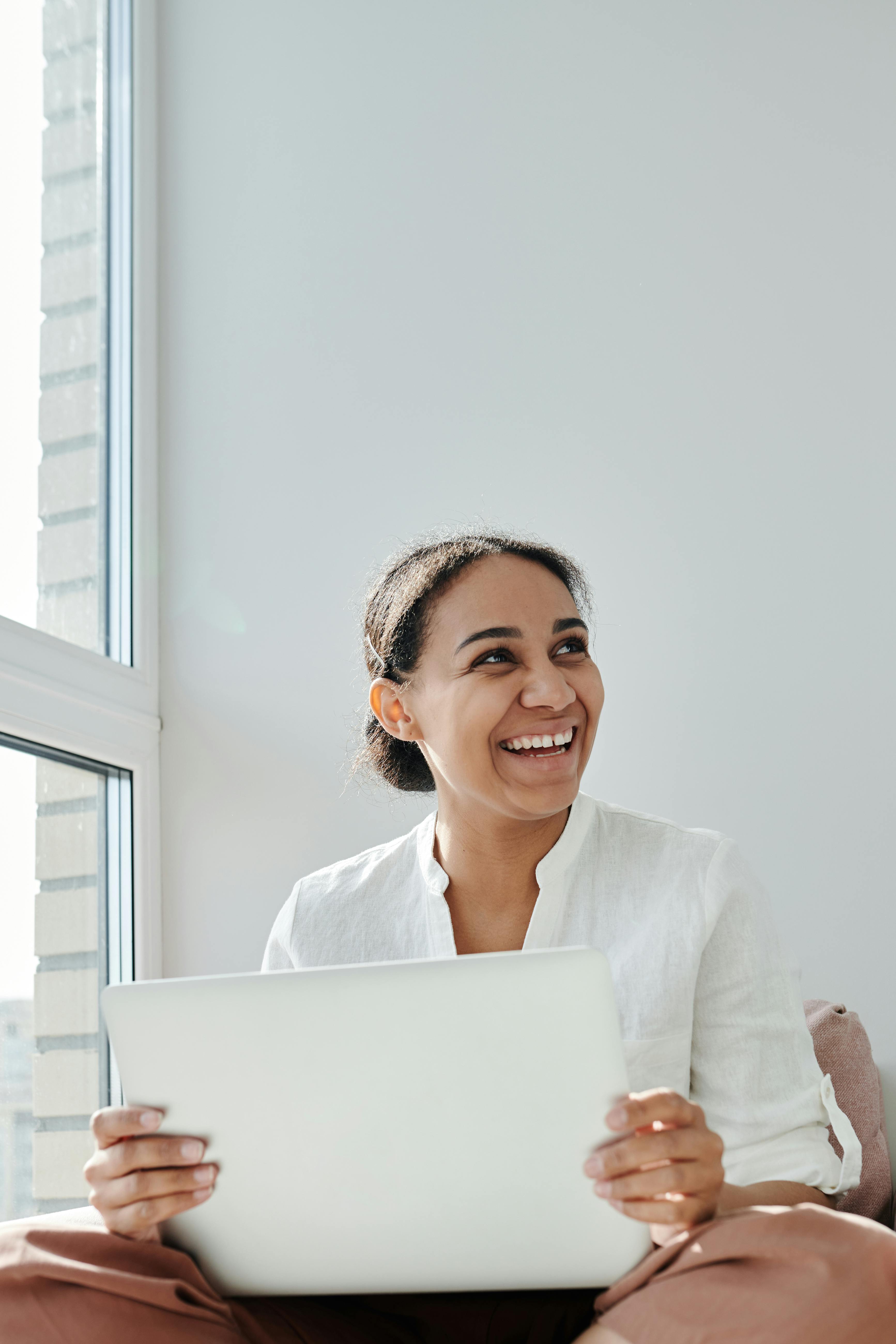 Smiling woman sitting by a window with a laptop, engaged in online work or study.