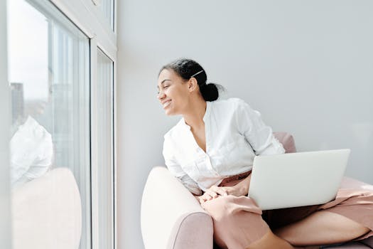 Woman enjoying a relaxing moment indoors, looking thoughtfully out the window with a laptop on her lap.