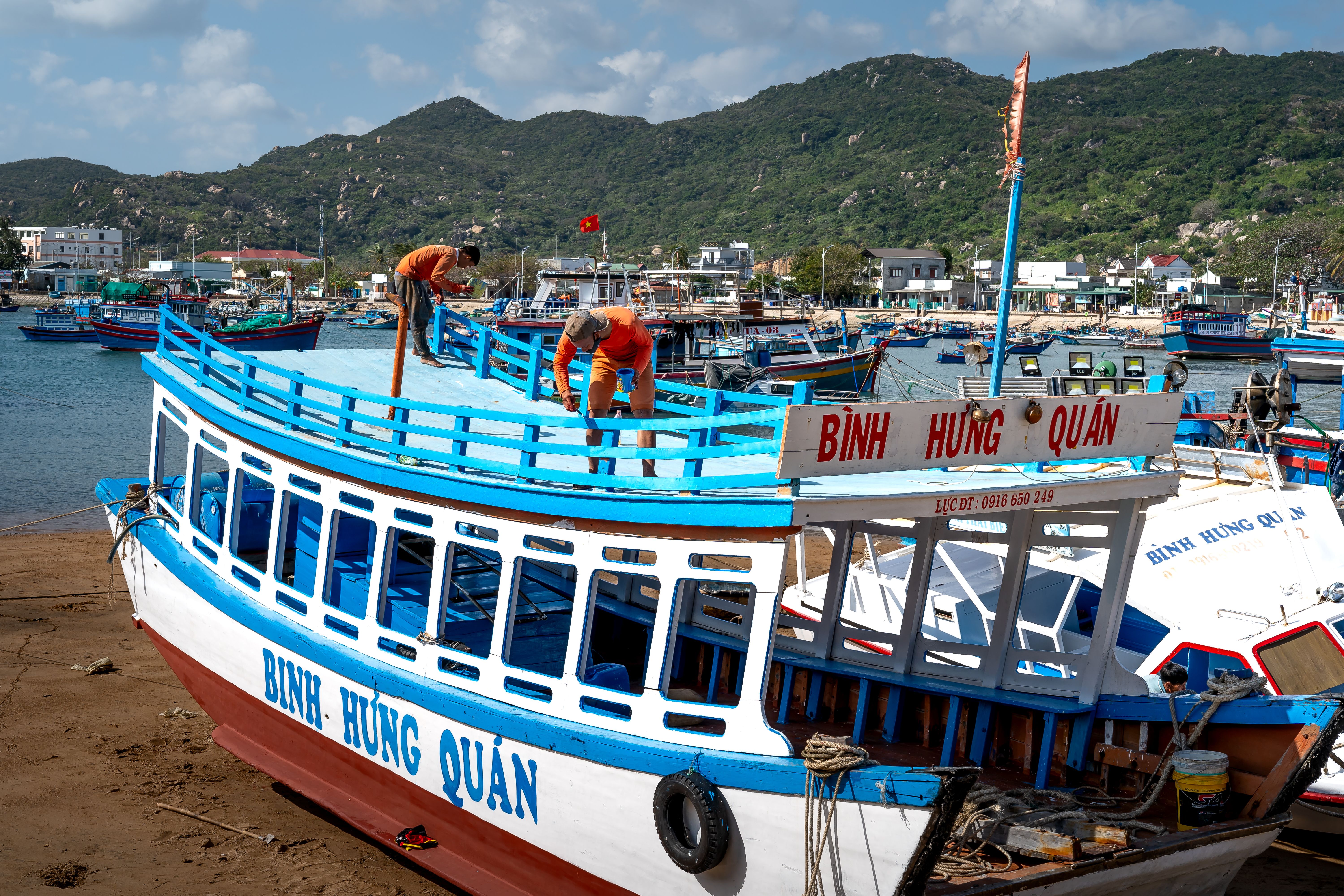 A traditional Vietnamese boat being repaired by workers in a bustling harbor setting with mountains in the background.