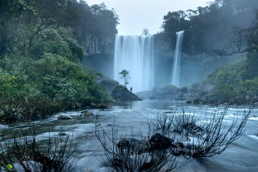 A lone figure stands by a majestic waterfall, surrounded by lush greenery and fog in a tranquil forest.
