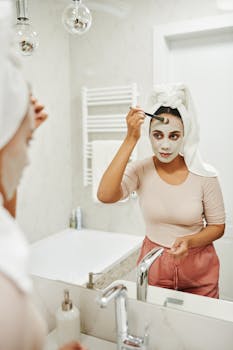 Woman applying clay mask with a brush in her bathroom mirror reflection.