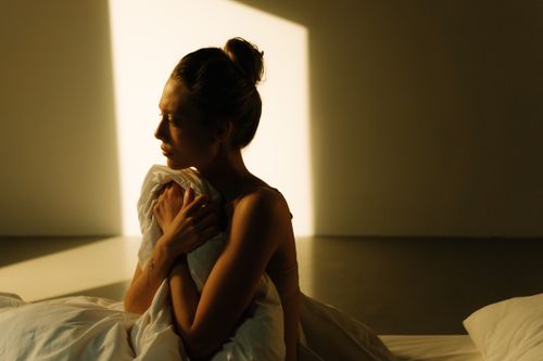 Free A woman sitting in soft morning light, wrapped in a blanket, exuding calmness. Stock Photo