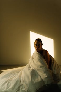 A person sits wrapped in a white blanket, illuminated by morning sunlight from a window.