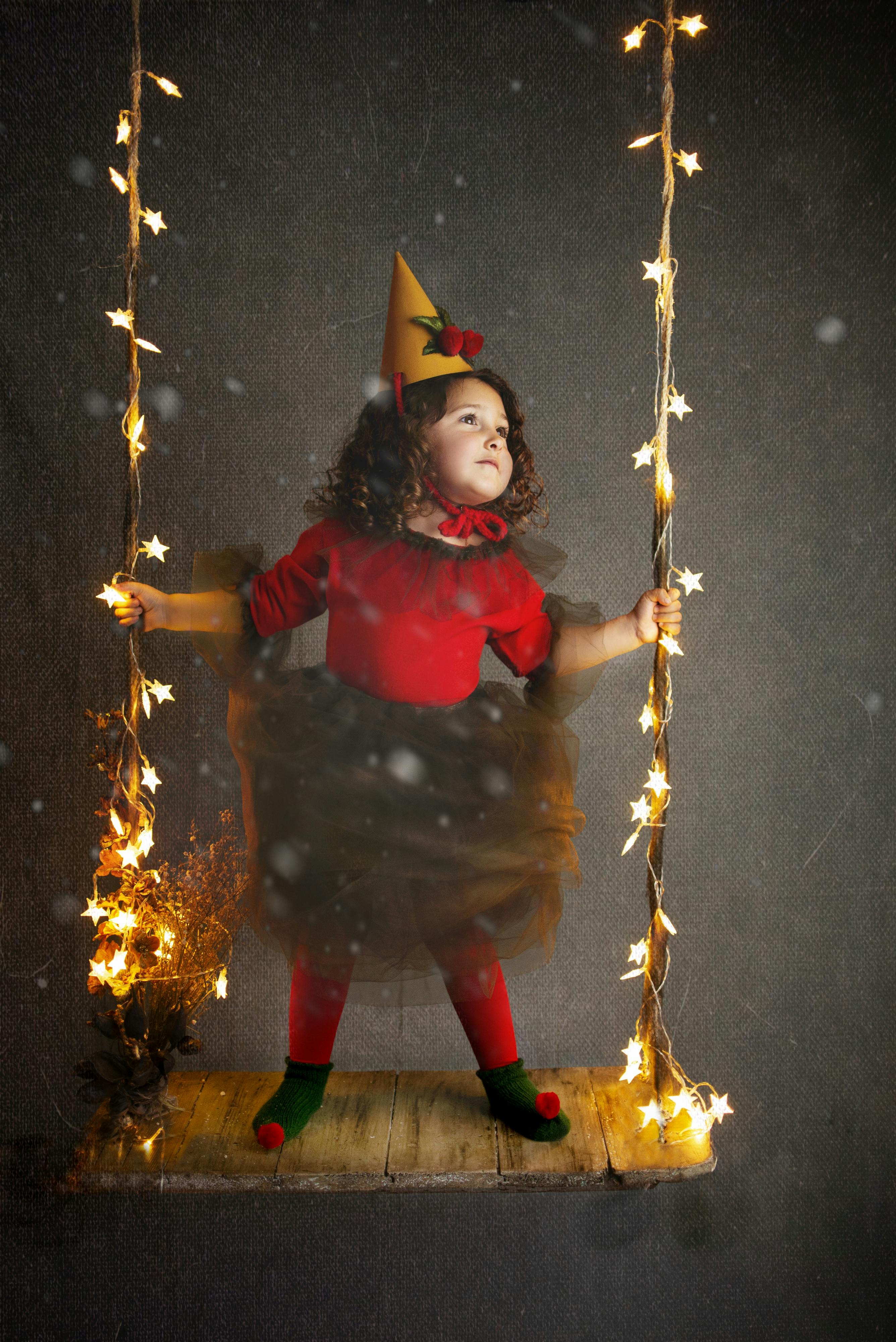 A young girl in festive costume standing on a decorated swing amidst soft lighting.
