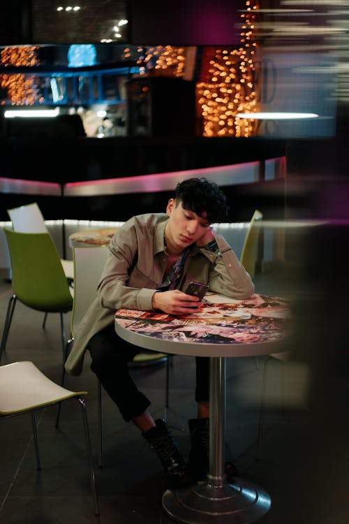 A Man in Beige Jacket Sitting Near the Table while Using His Mobile Phone
