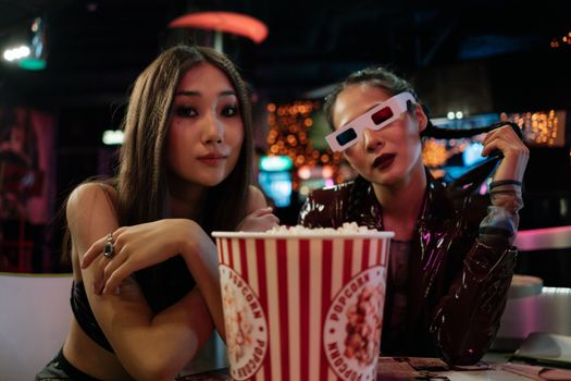 Two women with popcorn at a dimly lit theater, one wearing 3D glasses.