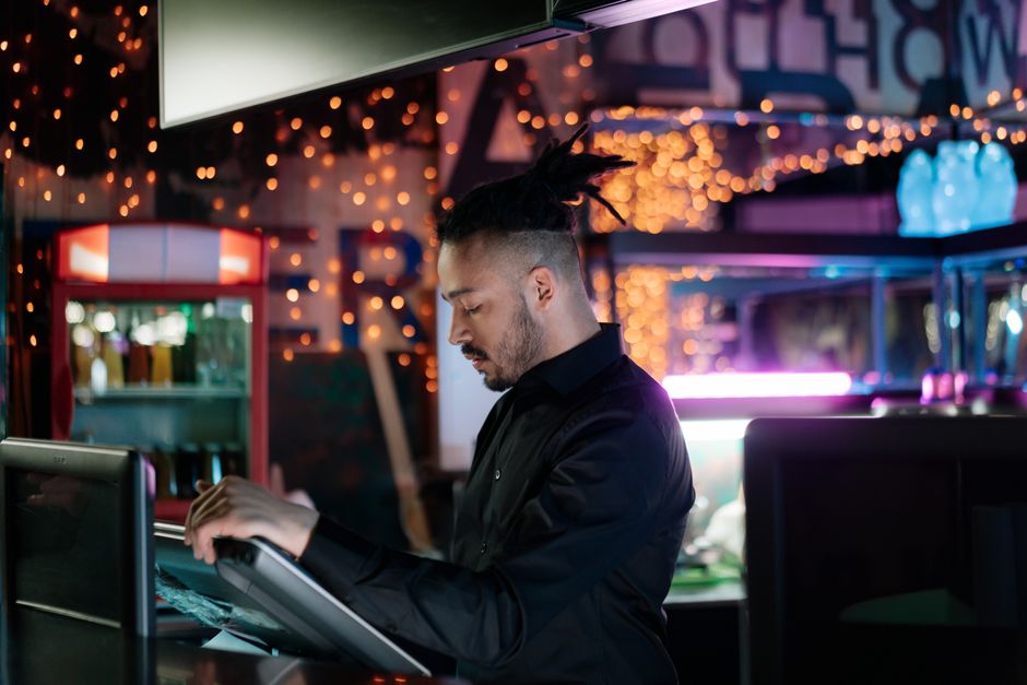 A man with facial hair operates a computer in a trendy bar with festive lights. A man with facial hair operates a computer in a trendy bar with festive lights.