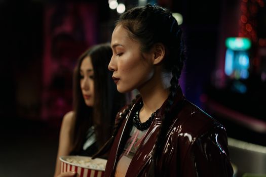 A fashionable Asian woman in a shiny jacket stands in a dimly lit venue, showcasing a side profile.