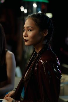 Profile portrait of an Asian woman in a leather jacket with braids, in moody lighting.