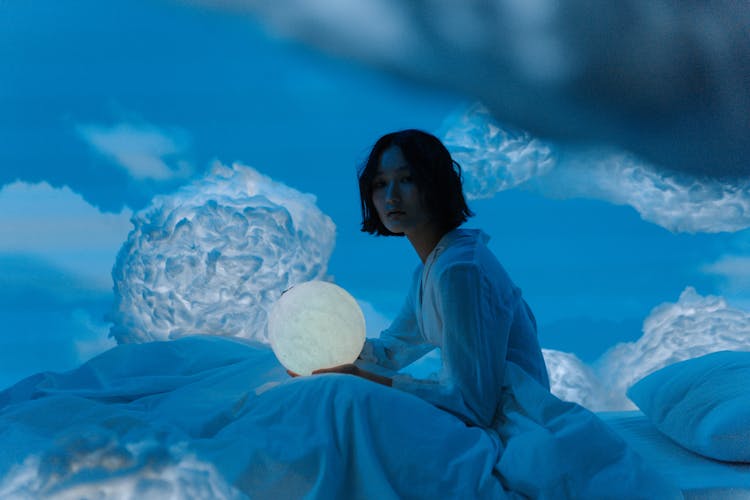 A Woman Sitting On The Bed While Holding A Moon Lamp 