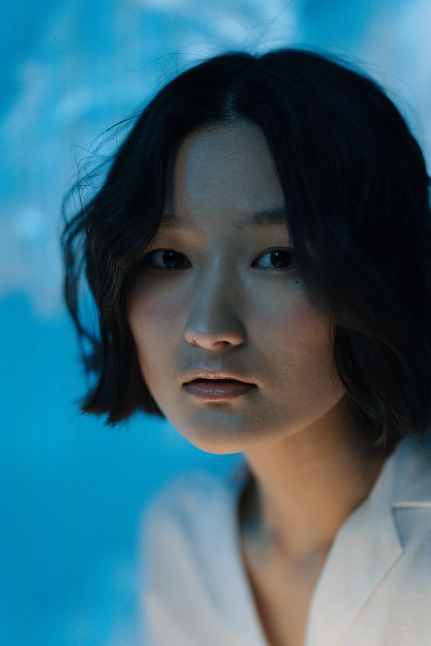Close-up portrait of an Asian woman indoors with a soft blue background, looking at the camera.
