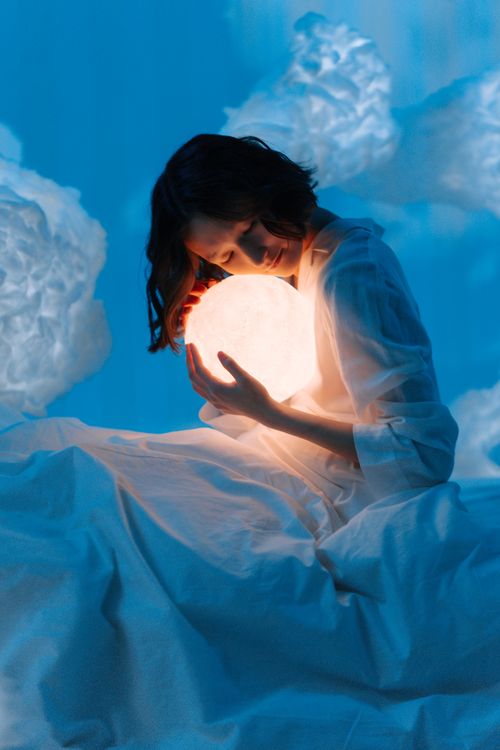 Free A woman in a surreal setting embraces a glowing sphere among soft cloud formations. Stock Photo