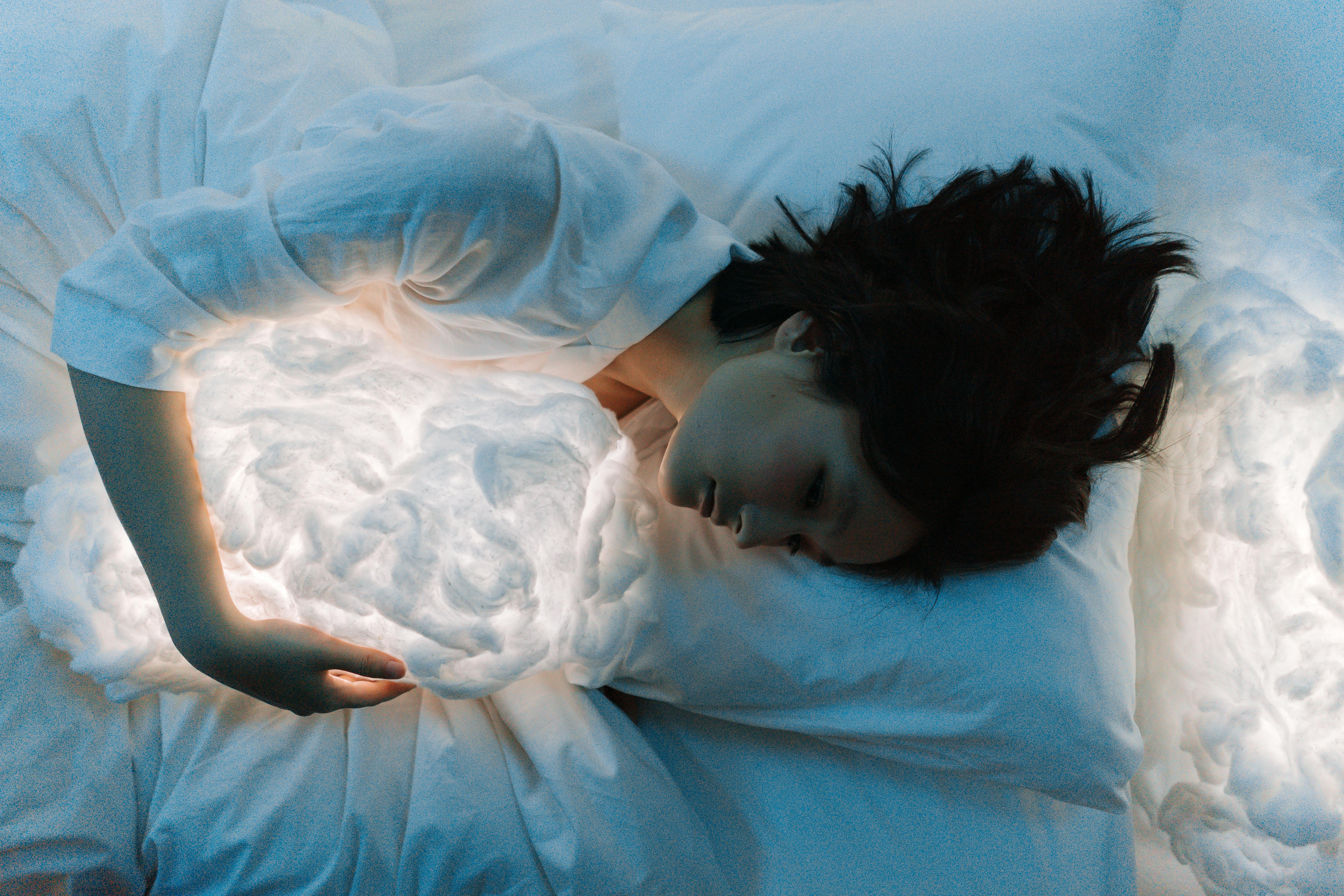 Woman in white long sleeves hugging a glowing pillow while resting in bed, creating a dreamlike atmosphere.