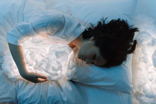 Free Woman in white long sleeves hugging a glowing pillow while resting in bed, creating a dreamlike atmosphere. Stock Photo