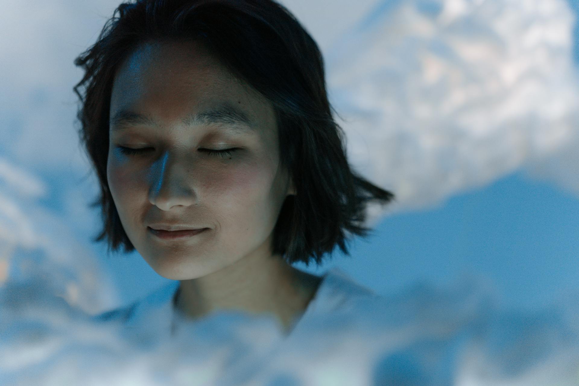 Peaceful portrait of a young Asian woman with closed eyes in a soft blue setting.