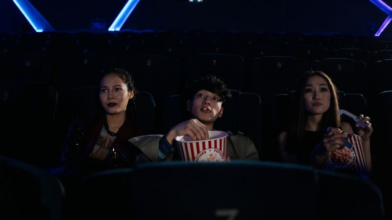 Three friends watching a movie in a theater, enjoying popcorn together.