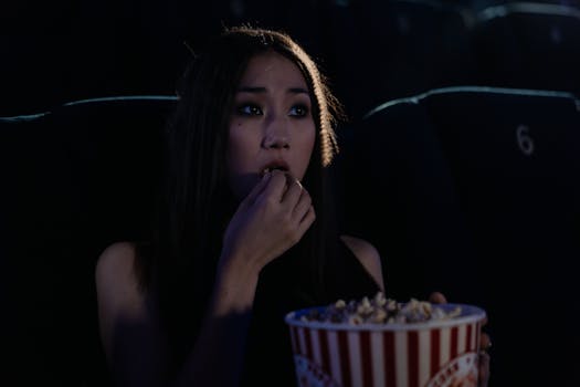 An East Asian woman enjoying popcorn while watching a movie in a theater.