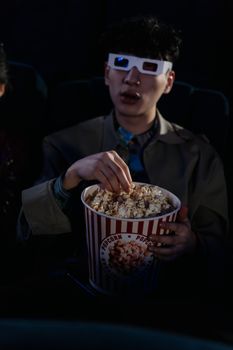 A young man wearing 3D glasses enjoys popcorn while watching a movie indoors.
