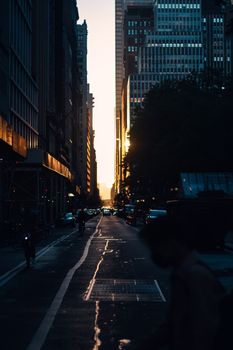 Anonymous people riding on motorcycle and pedestrian crossing asphalt road on narrow urban street with high buildings while dramatic sundown